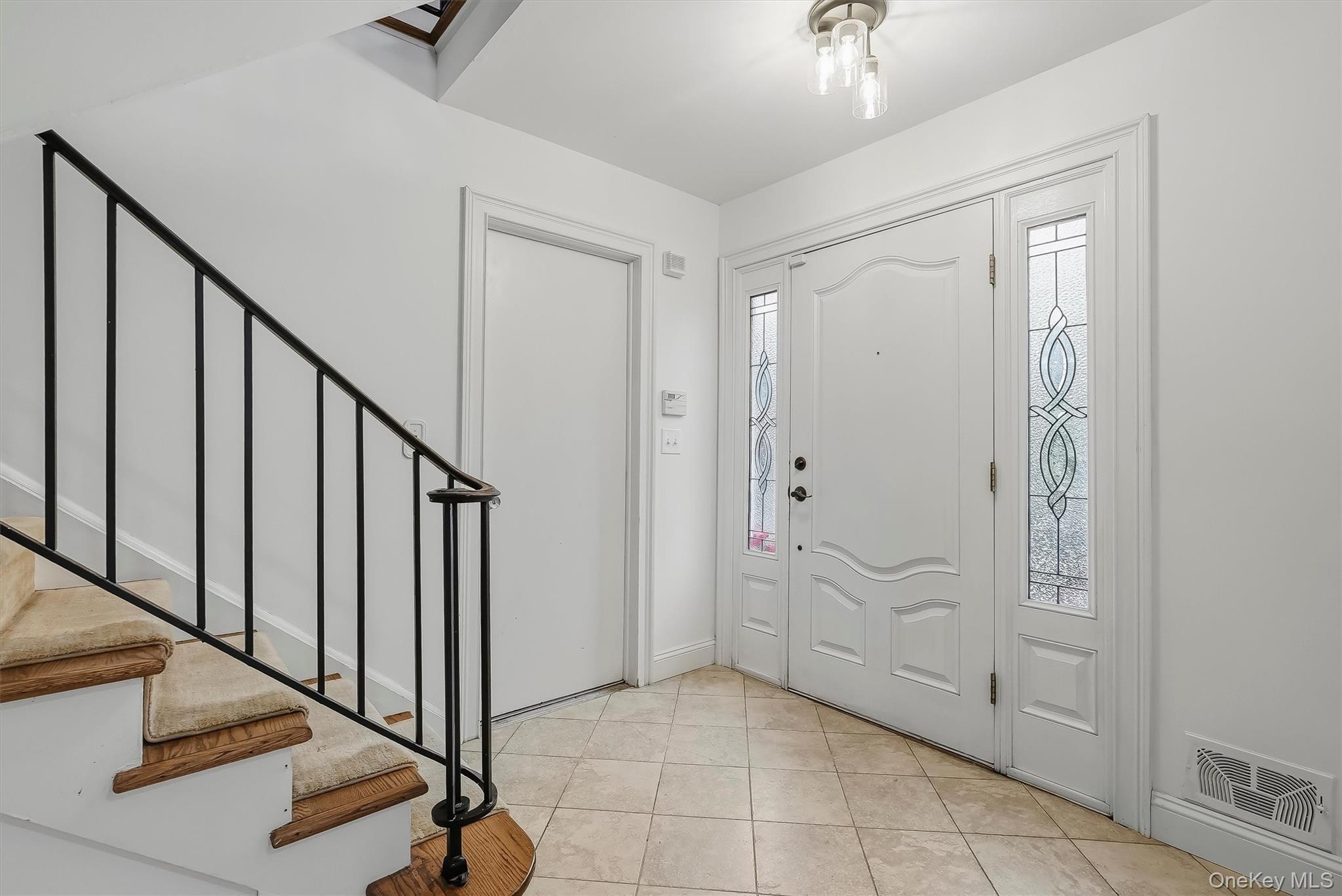 Front entry foyer with decorative glass sidelights, beige tile flooring, and a carpeted staircase with black railing.