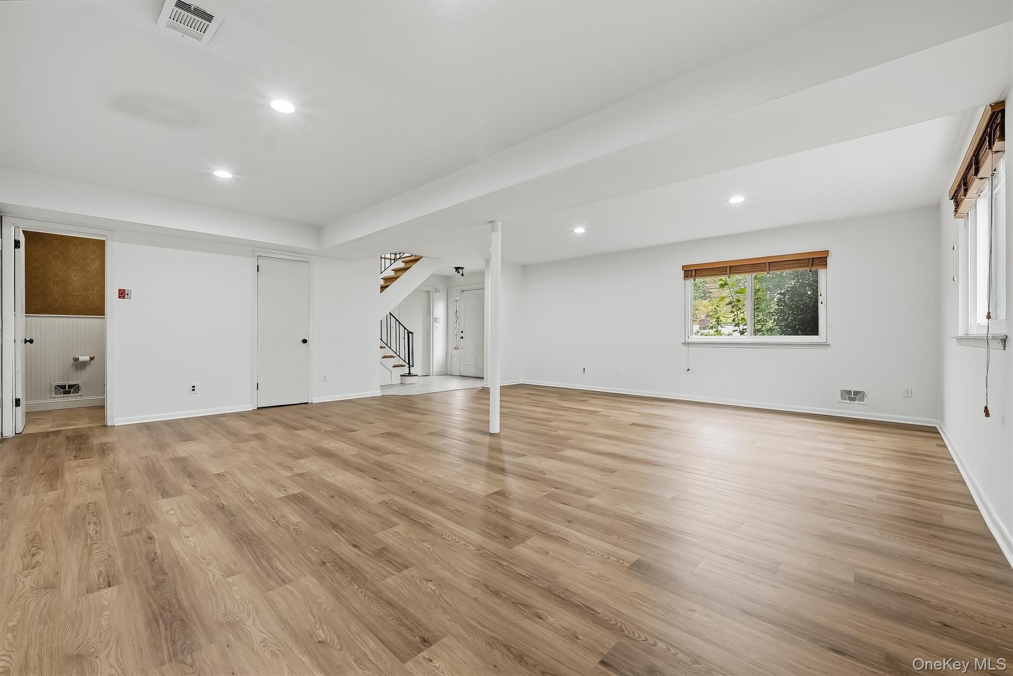 Empty living area with light wood-look floors, recessed lights, and a staircase at the back.