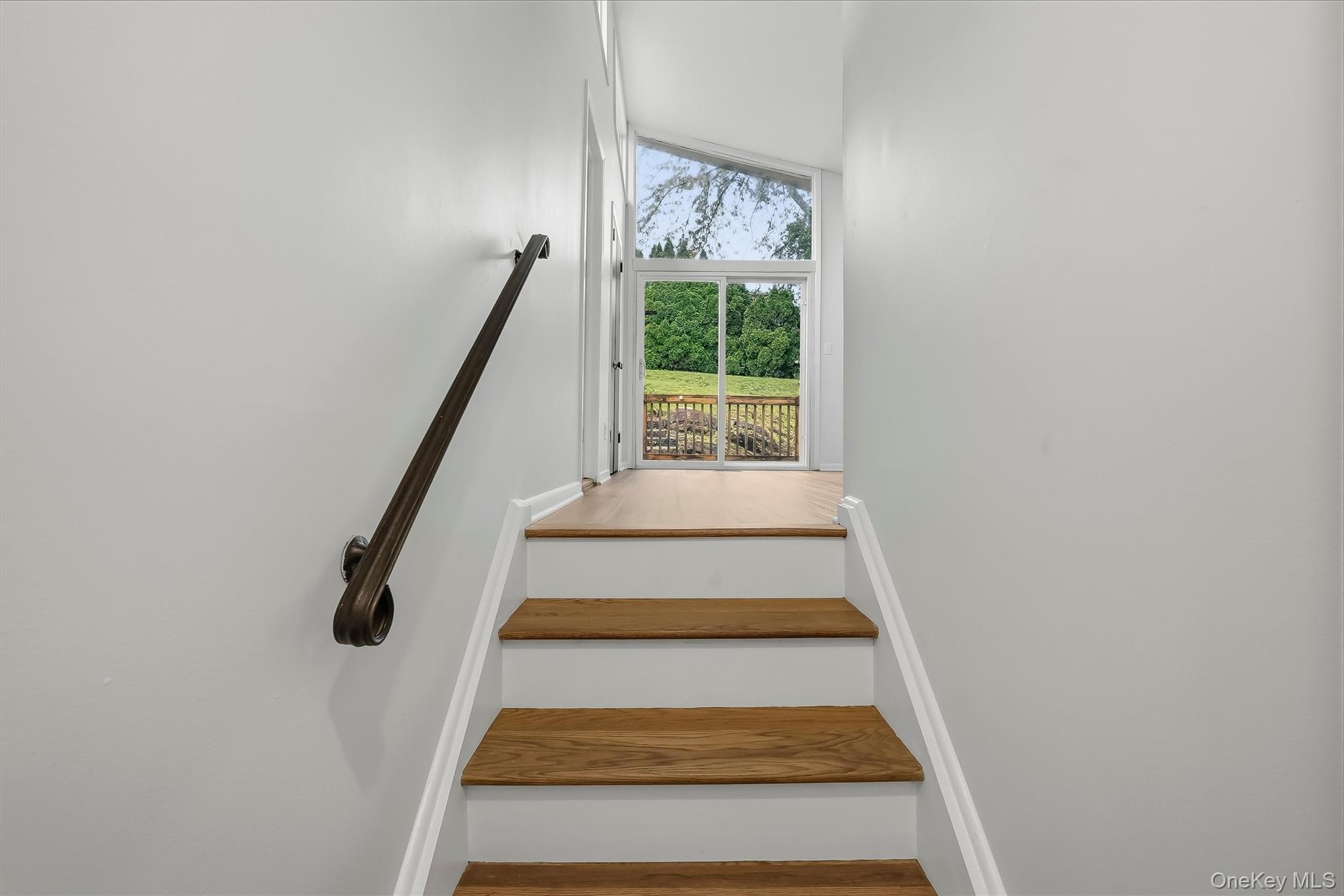 Wood-tread staircase with a dark handrail leads to a bright landing with a glass door overlooking a green yard.
