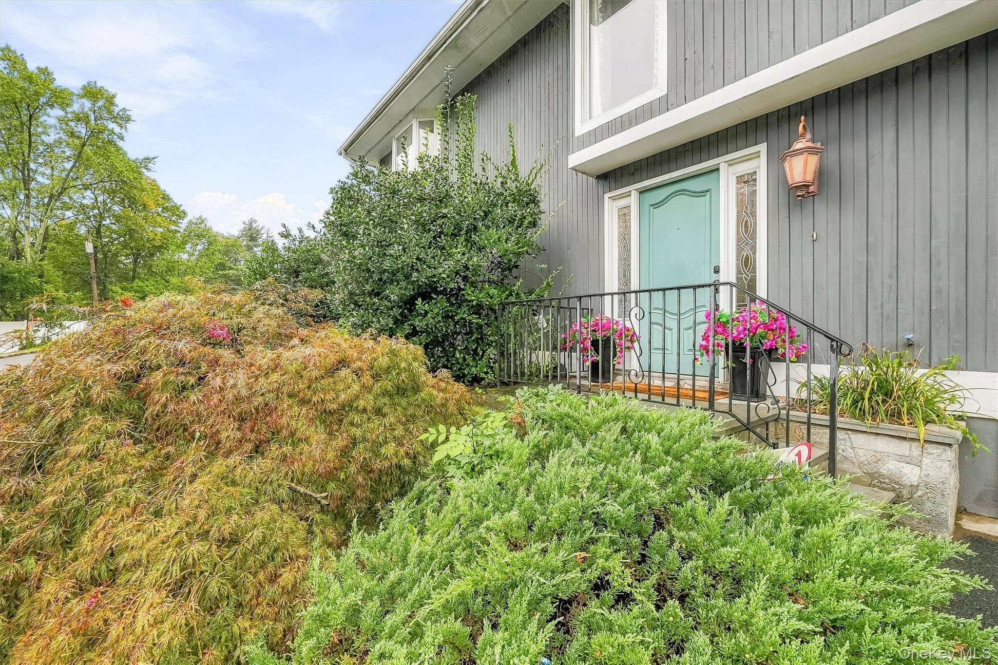 Gray house entrance with a turquoise door, black railing, pink flowers, and dense shrubs.