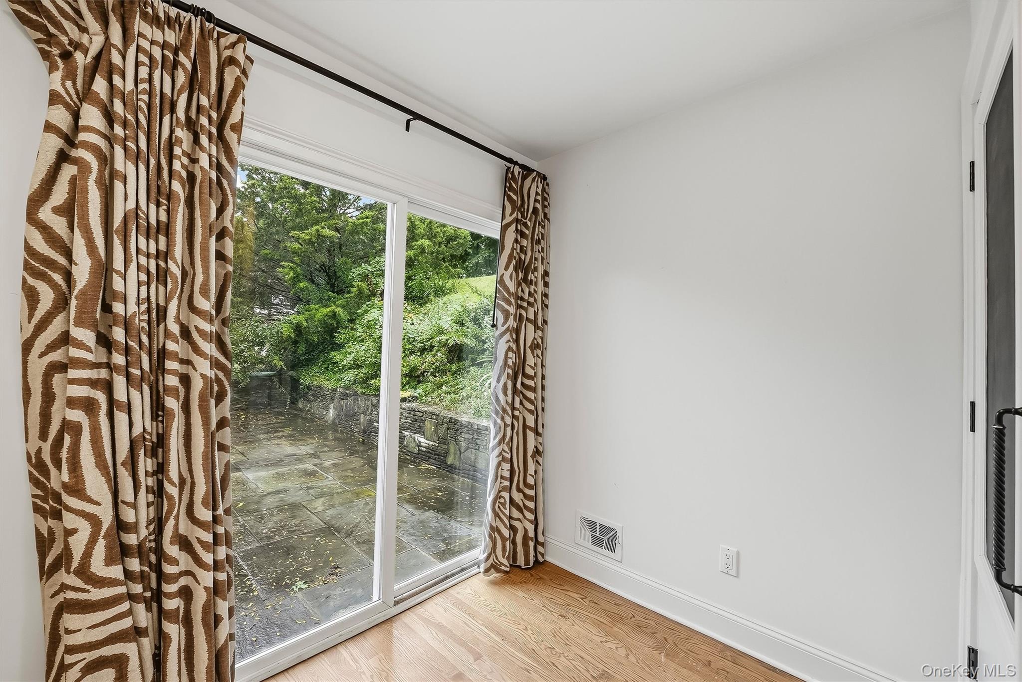 Unfurnished room with hardwood floors, patterned curtains, and a sliding glass door to a stone patio.
