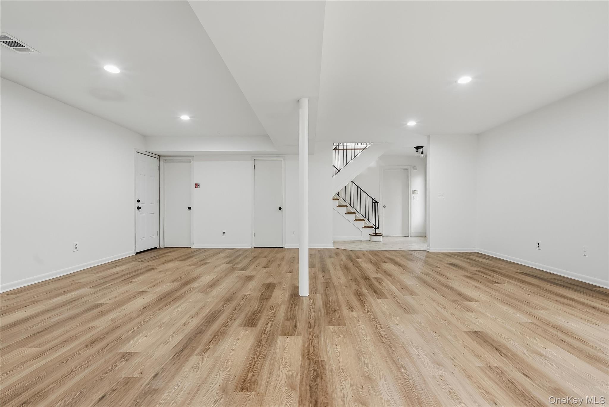 Empty lower-level room with wood-look flooring, a central support column, and a staircase.