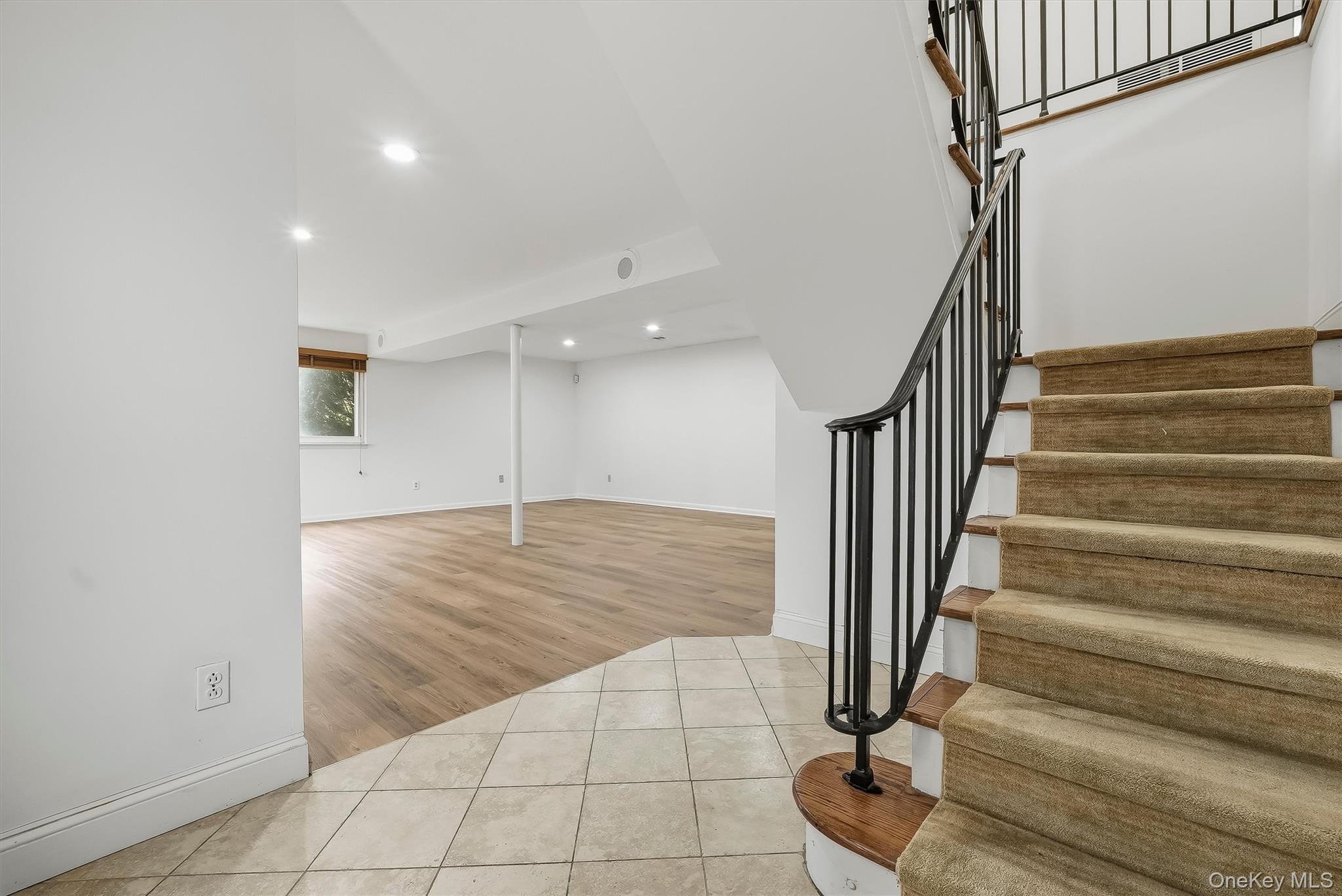 Empty lower-level room with light wood floors beside a carpeted staircase with black railings.