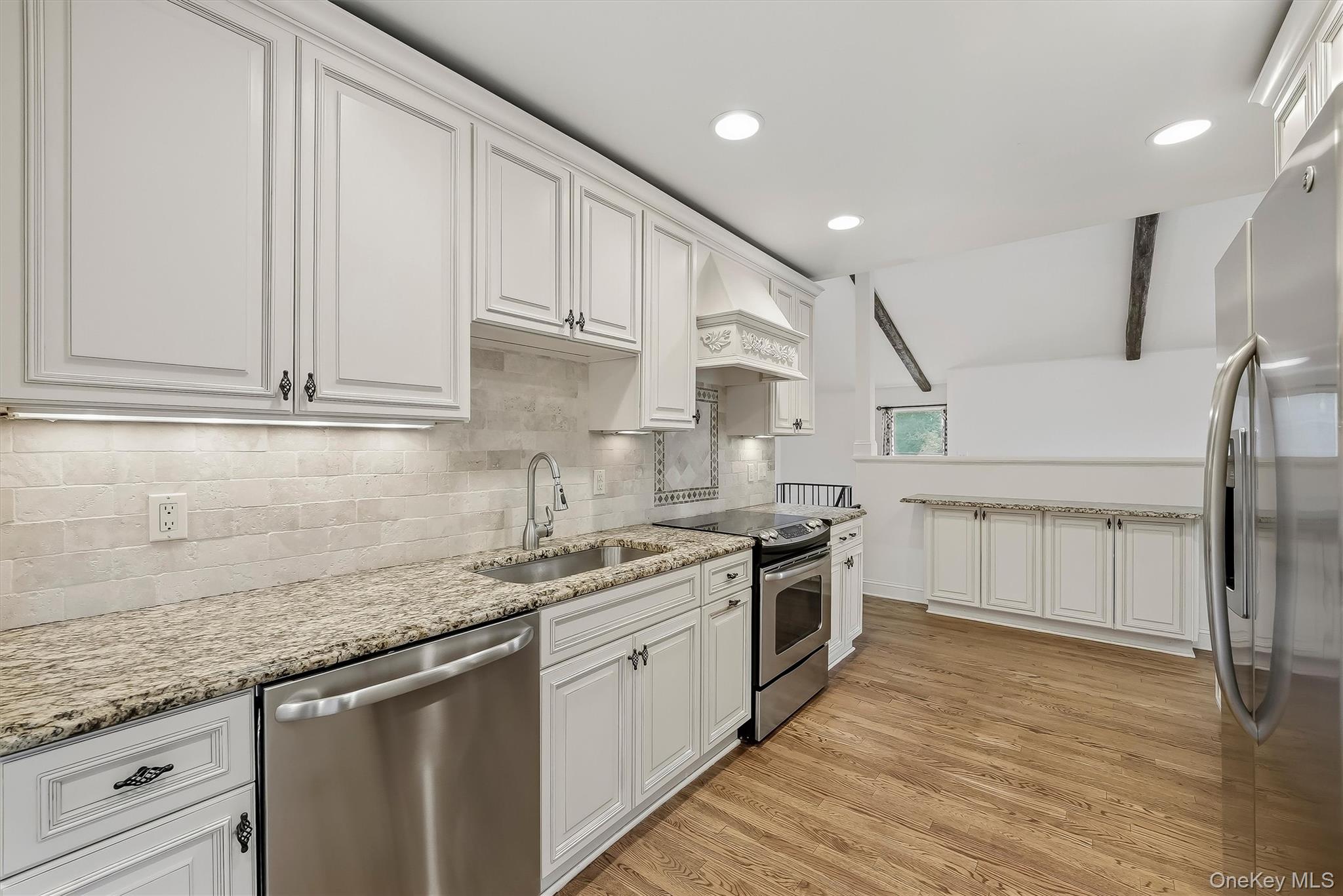 Kitchen with white paneled cabinets, granite counters, stainless appliances, hardwood floors, and exposed ceiling beams.