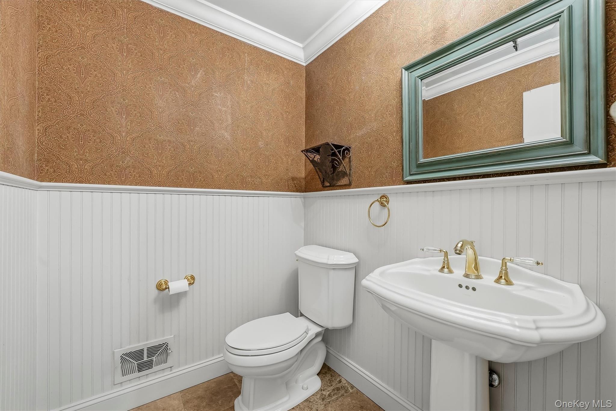 Bathroom with a pedestal sink, toilet, white beadboard wainscoting, and patterned tan wallpaper.