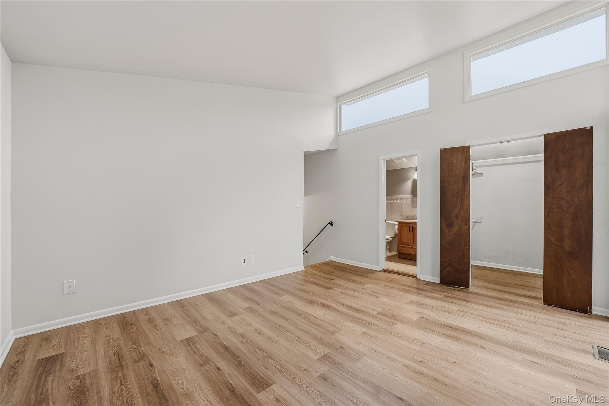 Empty bedroom with wood flooring, high windows, and sliding-door closet beside an adjoining bathroom.