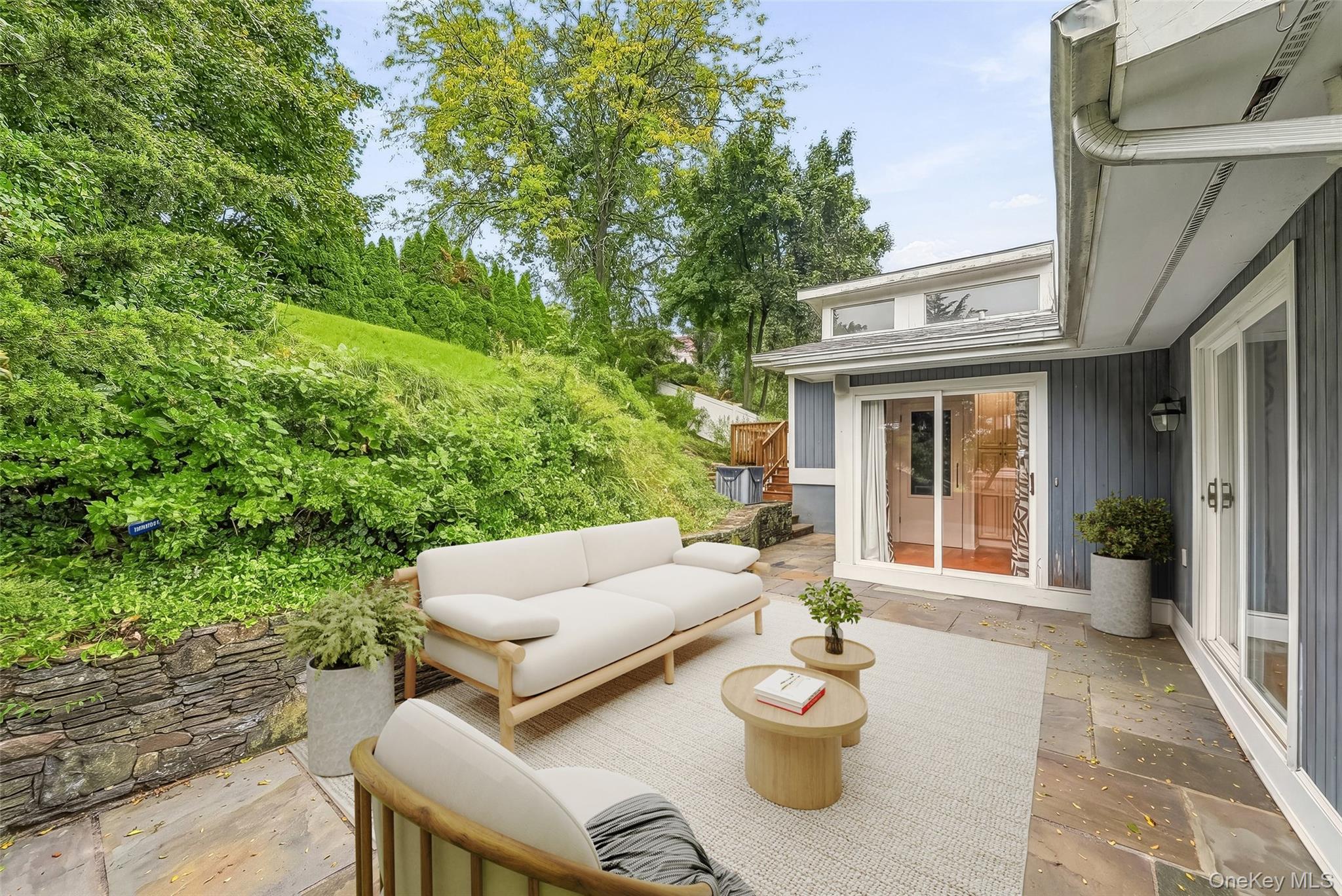 Staged patio with white outdoor seating, sliding glass doors, and a leafy hillside.