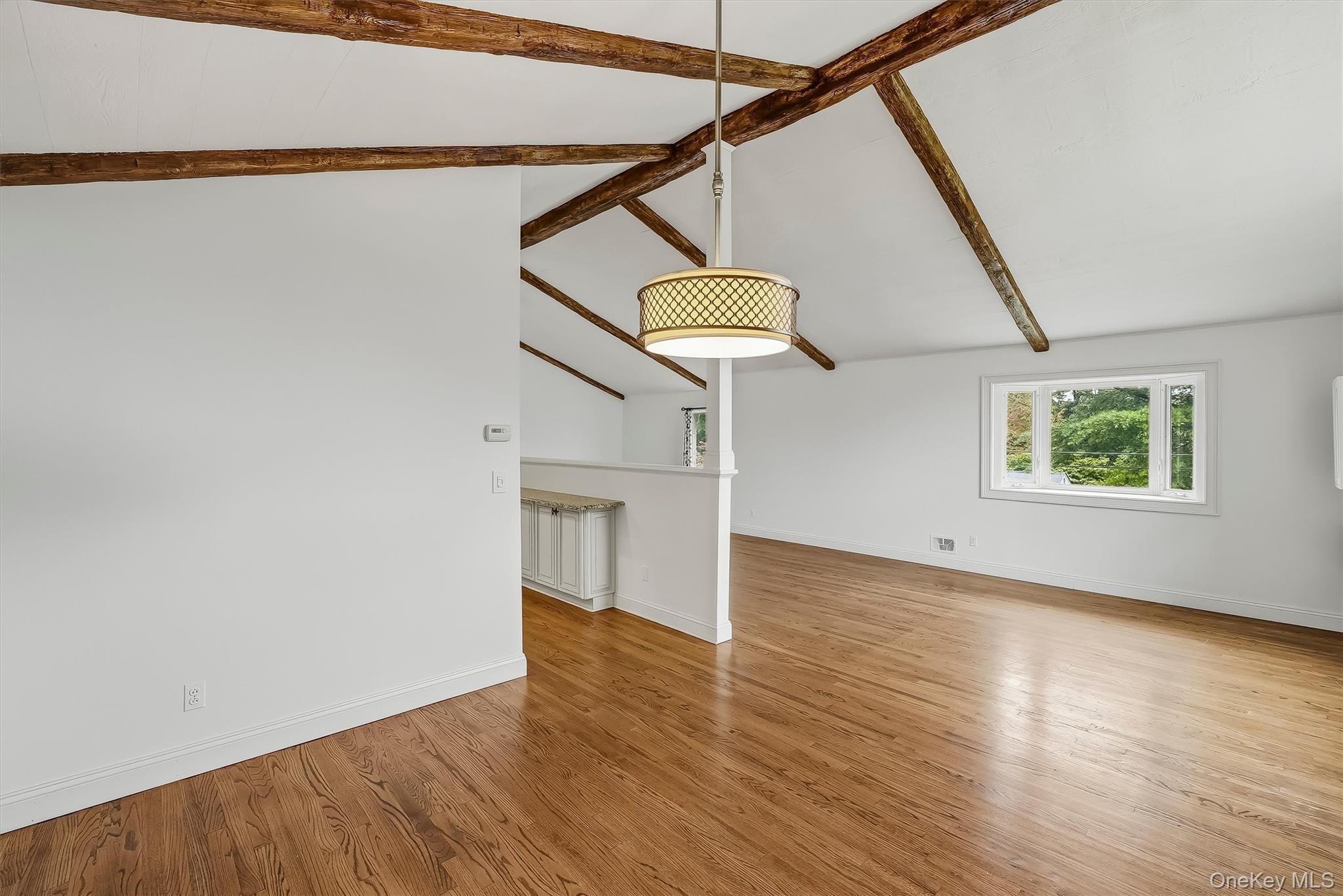 Open living area with exposed wood beams, hardwood floors, and a window overlooking greenery.
