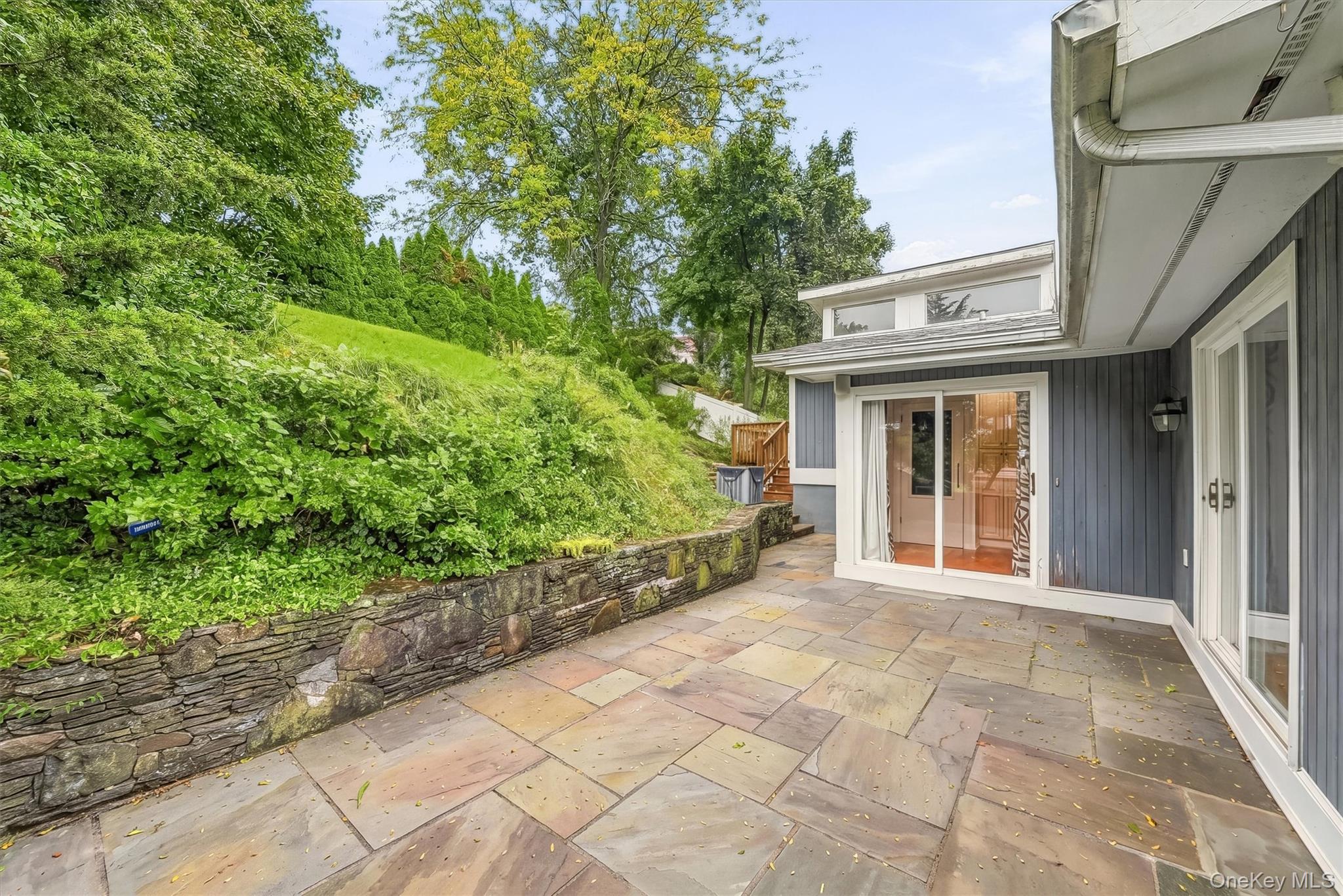 Flagstone patio beside a gray-sided home, bordered by a stone wall and lush sloping garden.