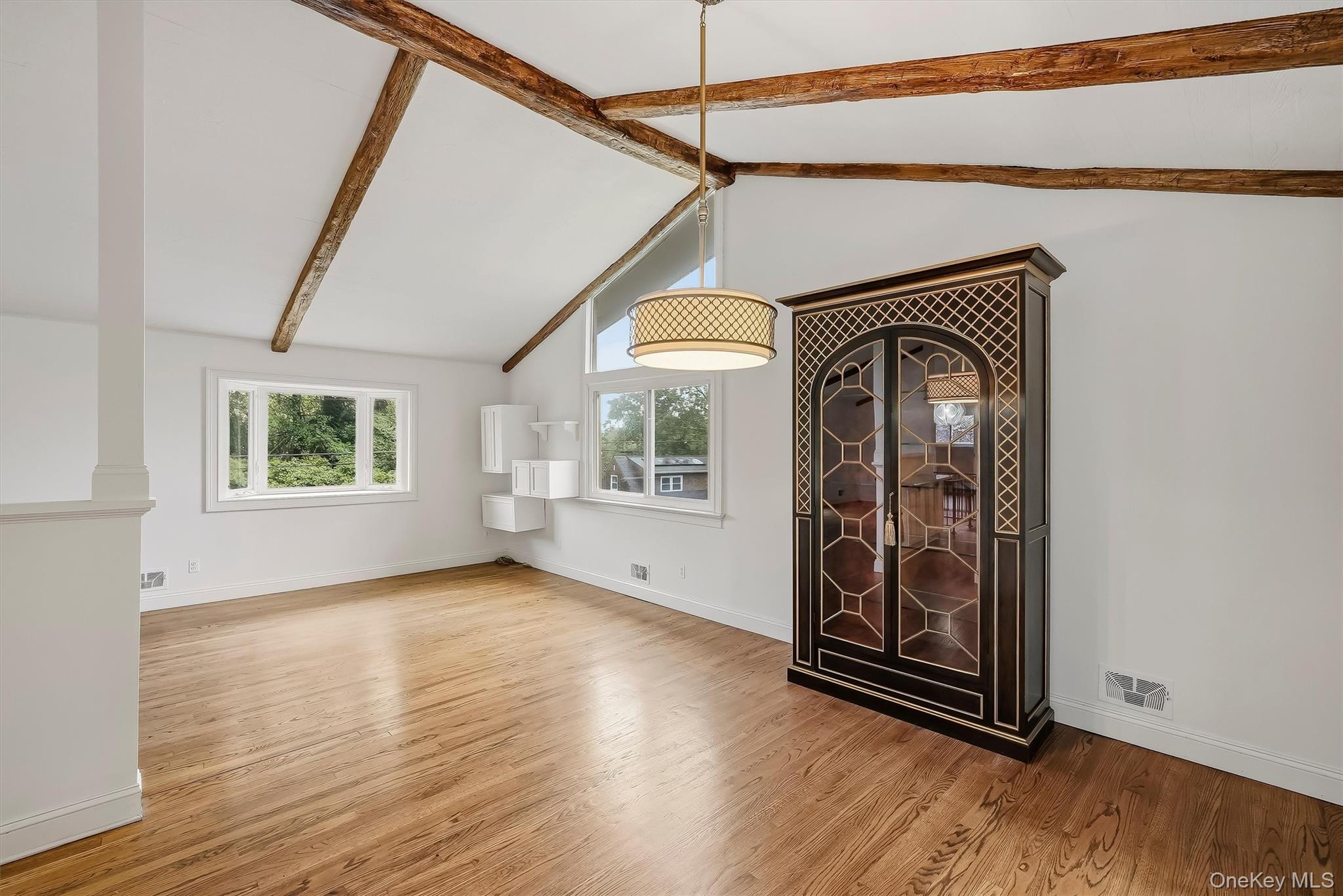 Bright room with hardwood floors, exposed ceiling beams, and a tall glass-fronted display cabinet.