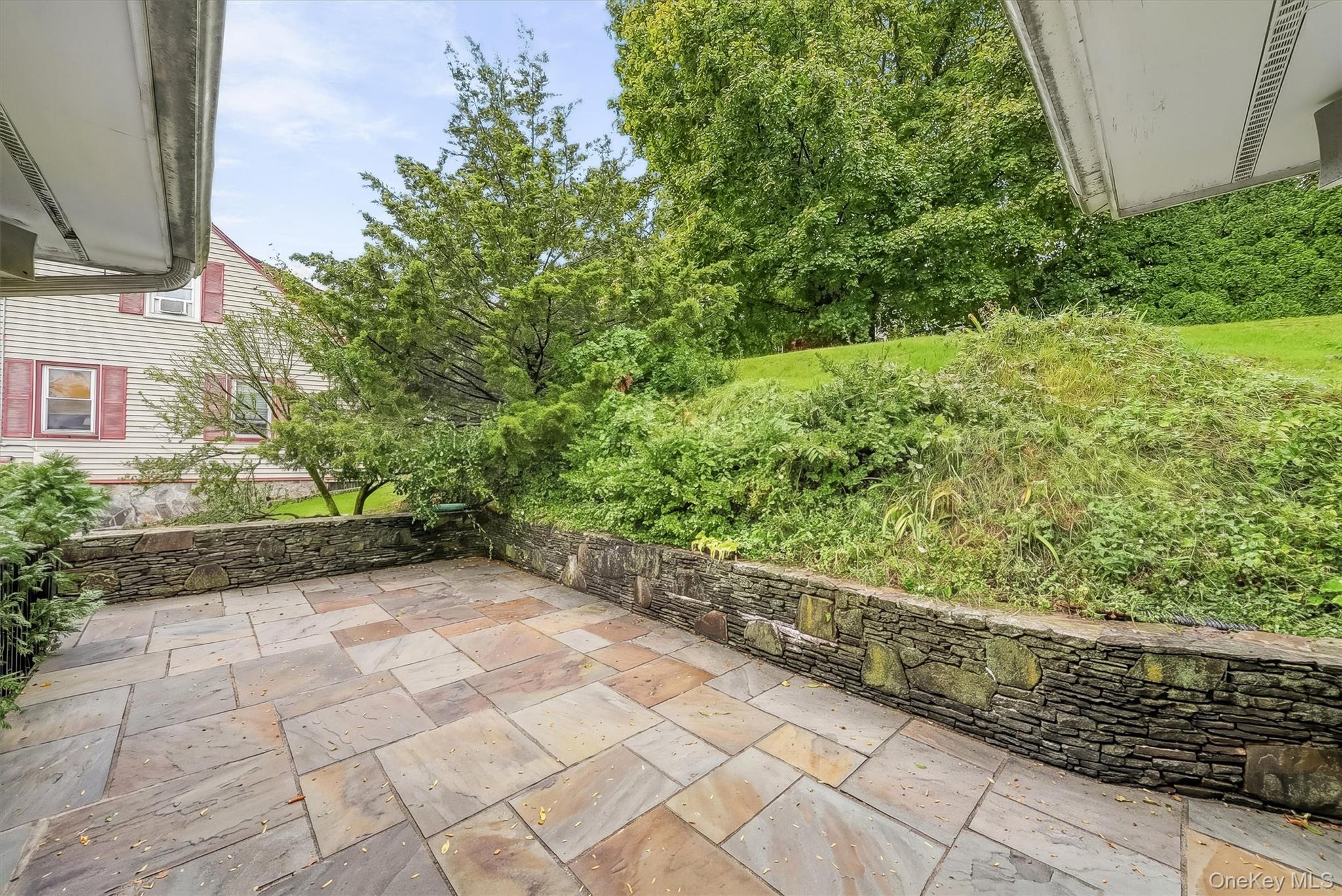 Stone-paved backyard patio bordered by stacked-stone walls, shrubs, and a grassy slope.