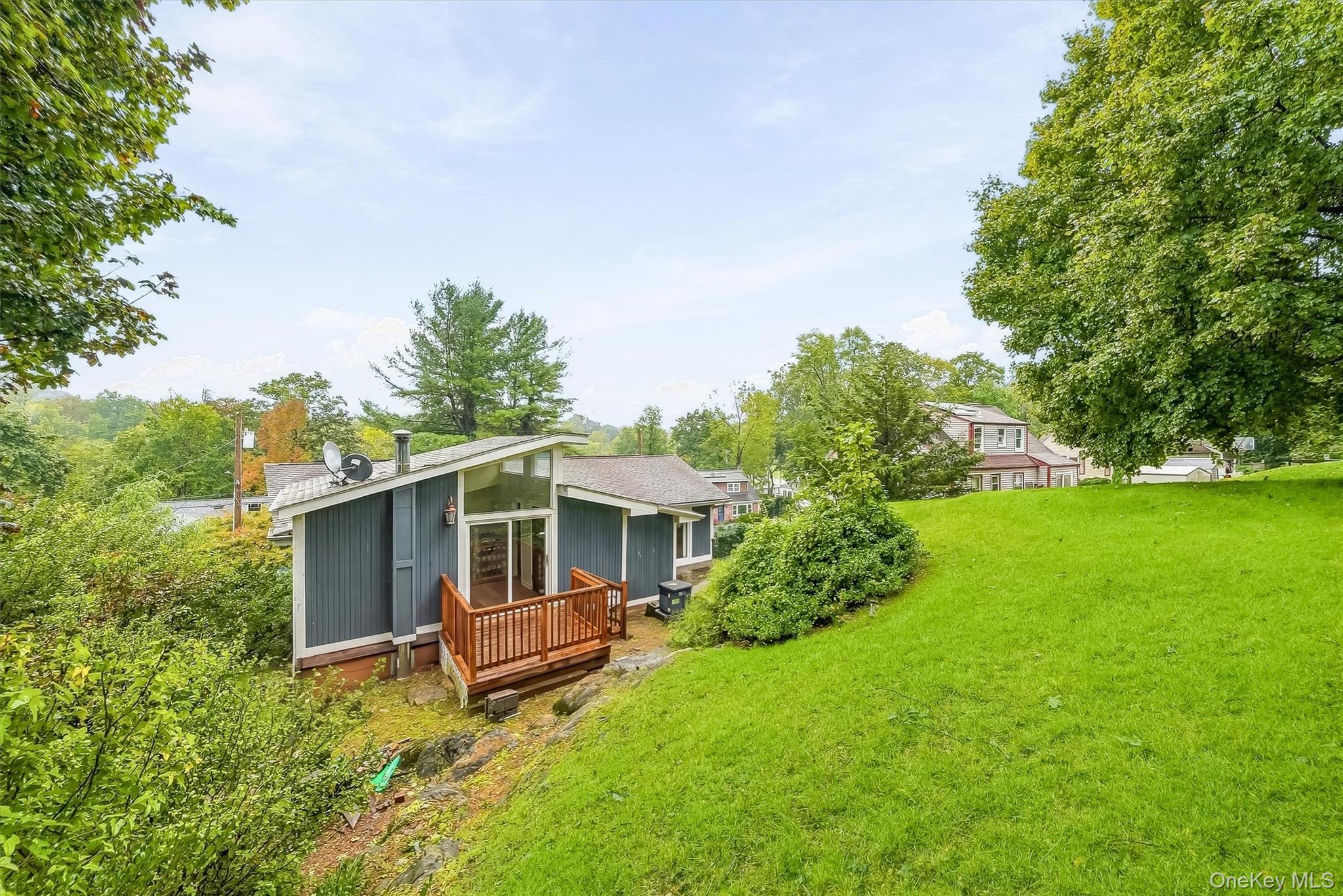 Blue-gray home with a wooden deck on a wooded slope above a broad green lawn.