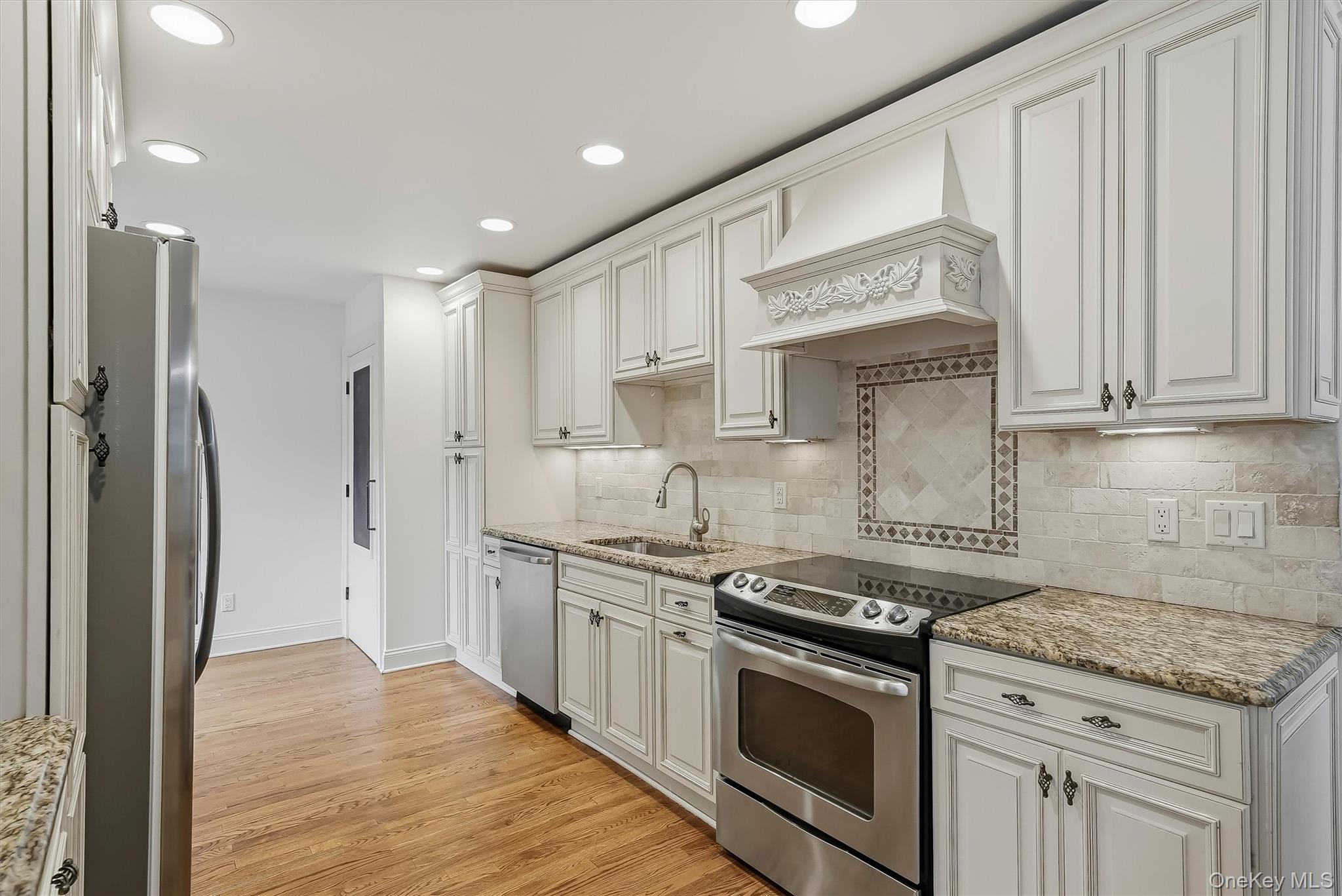 Kitchen with white cabinetry, stainless steel appliances, decorative range hood, and hardwood floors.