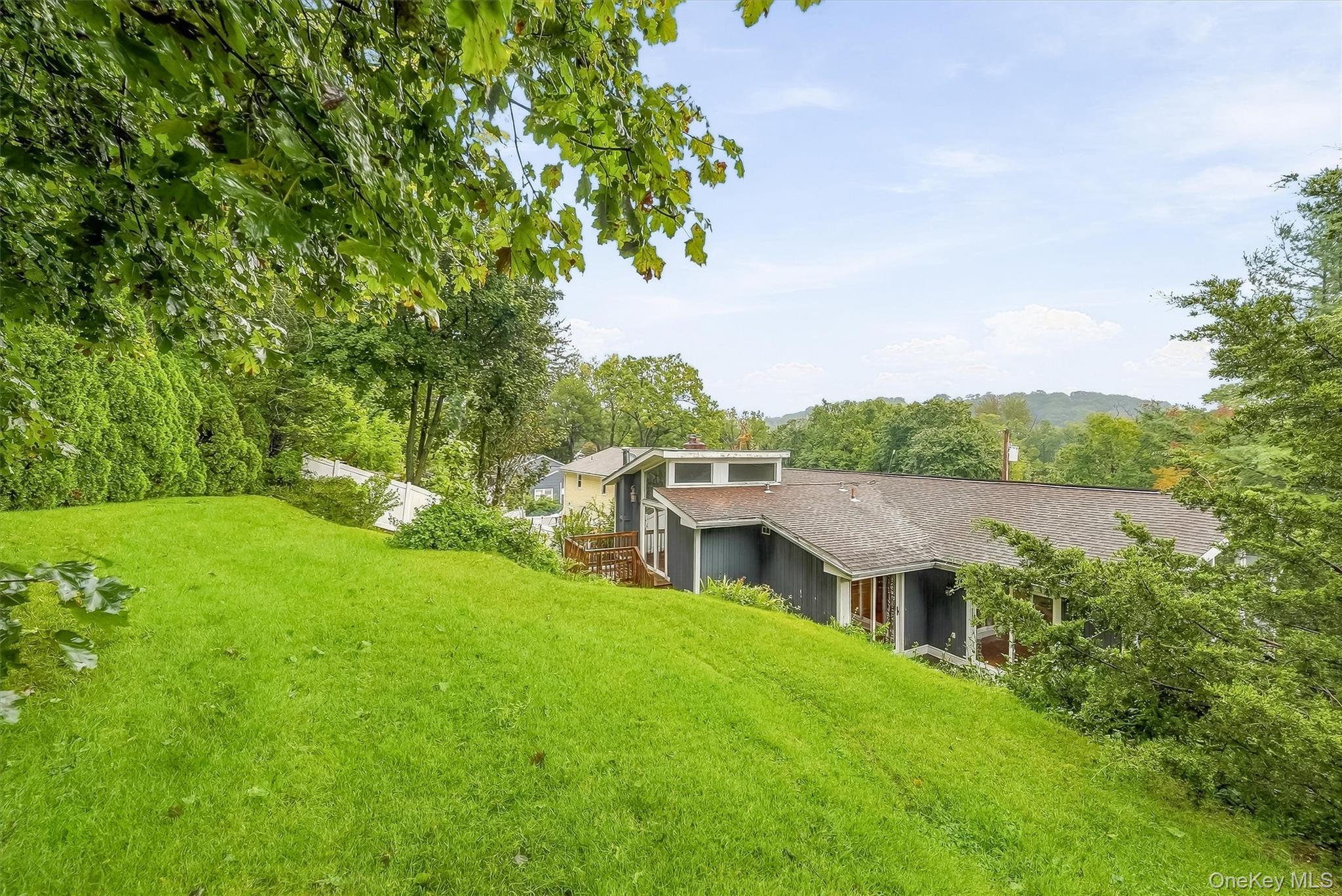 Low, dark-sided home set below a sloping lawn, surrounded by mature trees and shrubs.