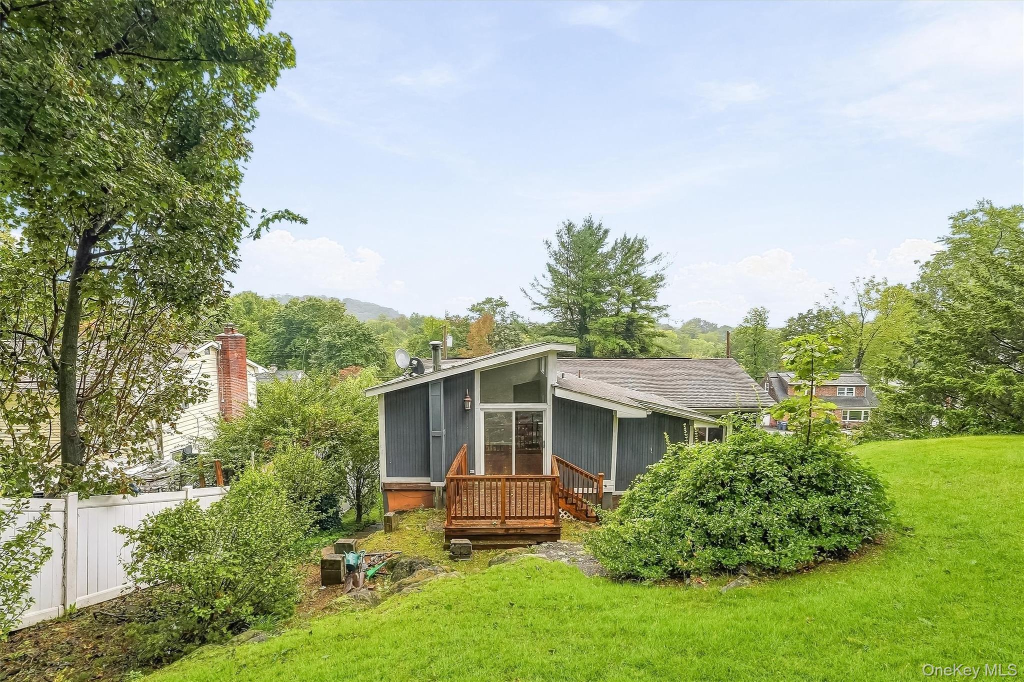 Rear exterior of a gray home with a wooden deck, sloping lawn, and mature trees.