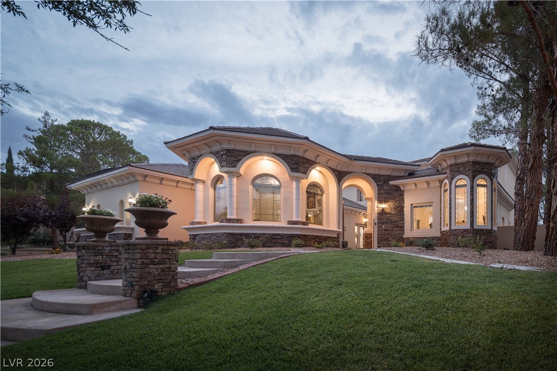 Luxury home exterior with illuminated arched windows, stone accents, and a landscaped lawn at dusk.