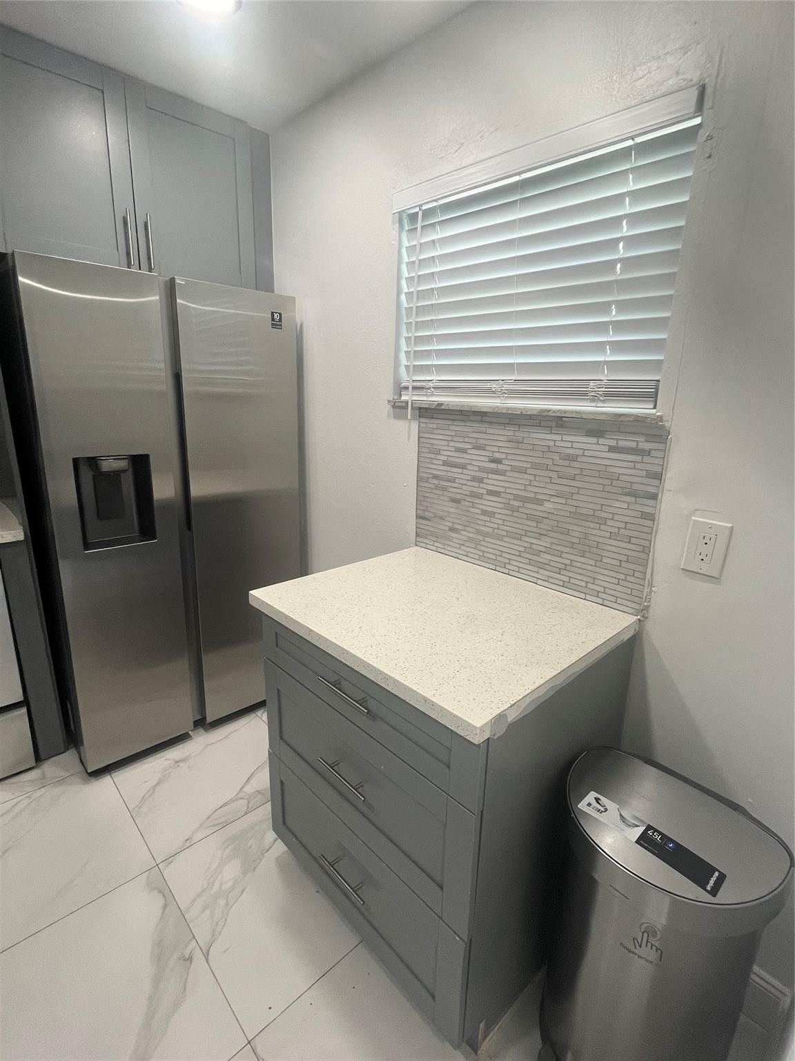 Kitchen with gray cabinetry, stainless steel refrigerator, white countertop, and tiled backsplash beneath a window.