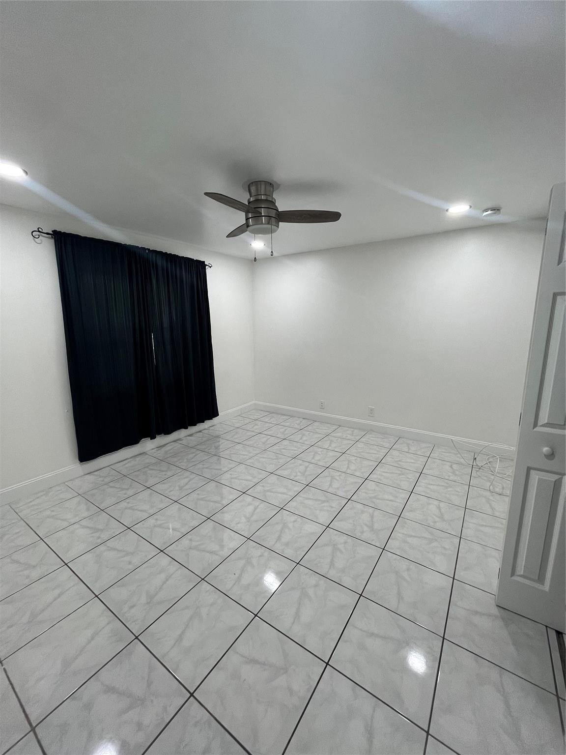 Empty room with marble-look tile floors, a ceiling fan, and dark window curtains.