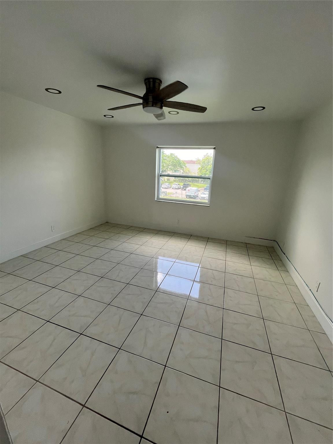 Empty room with tiled floors, a dark ceiling fan, and a window overlooking a parking area.
