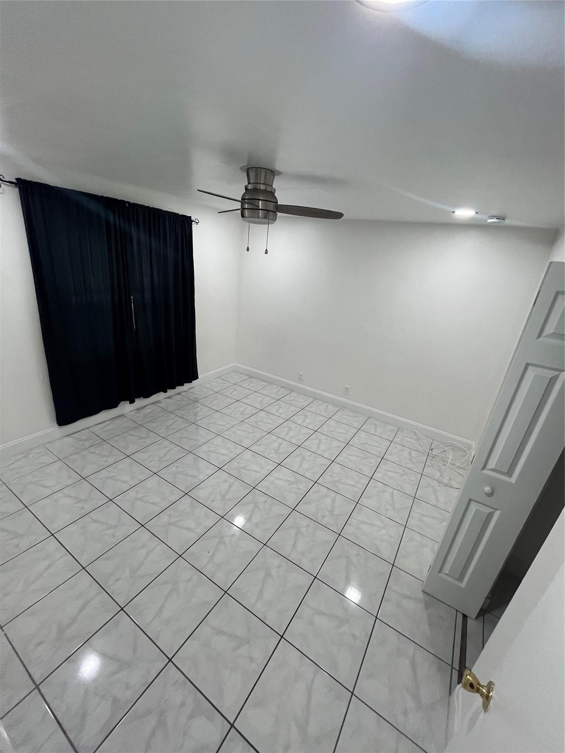 Empty tiled room with black curtains, a ceiling fan, and an open paneled door.