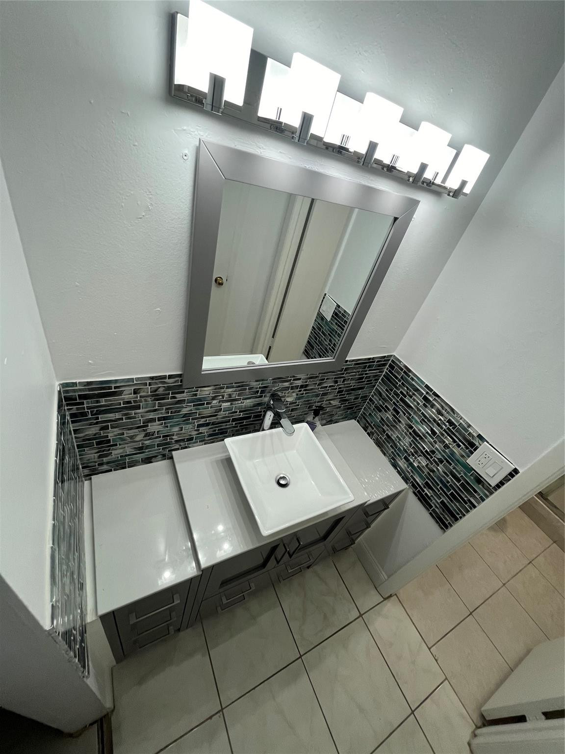 Bathroom vanity with a white sink, mosaic tile backsplash, large mirror, and bright lights.