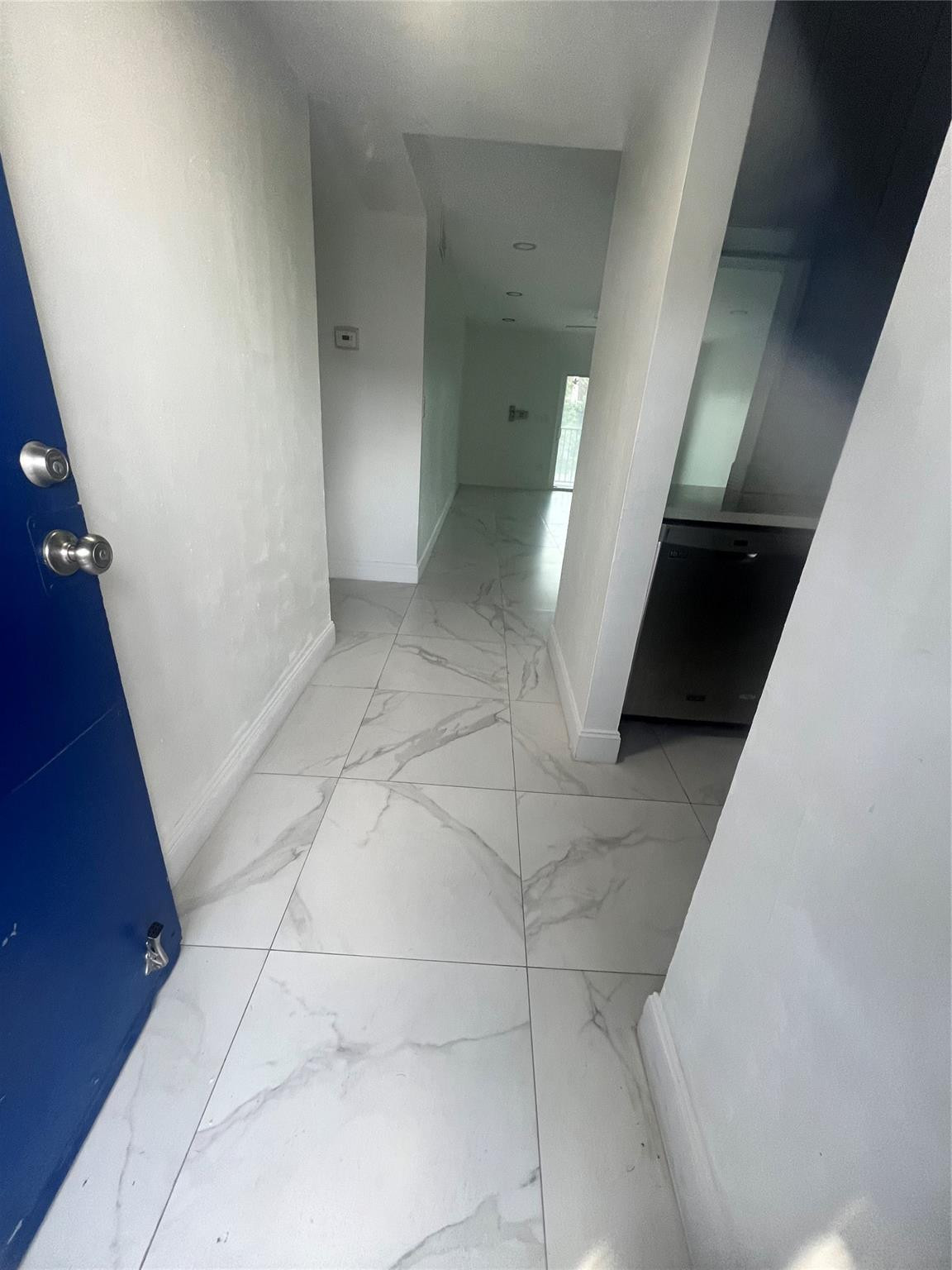 Unfurnished hallway with marble-look tile floors, an open blue door, and a bright window at the far end.