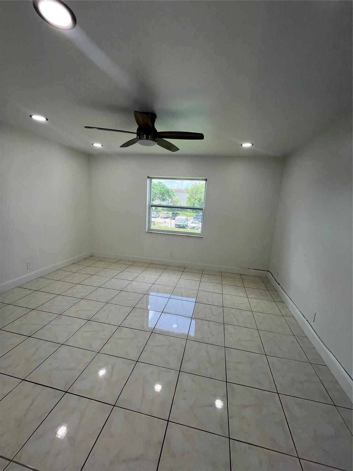 Empty tiled room with a ceiling fan and a window overlooking a parking area.