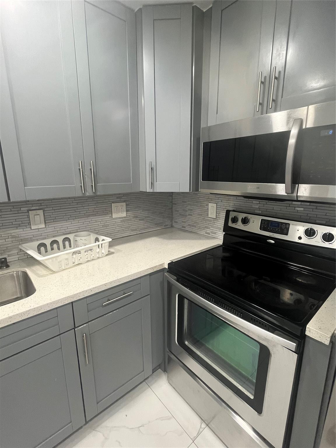 Modern kitchen with gray cabinets, light counters, mosaic backsplash, and stainless steel appliances.