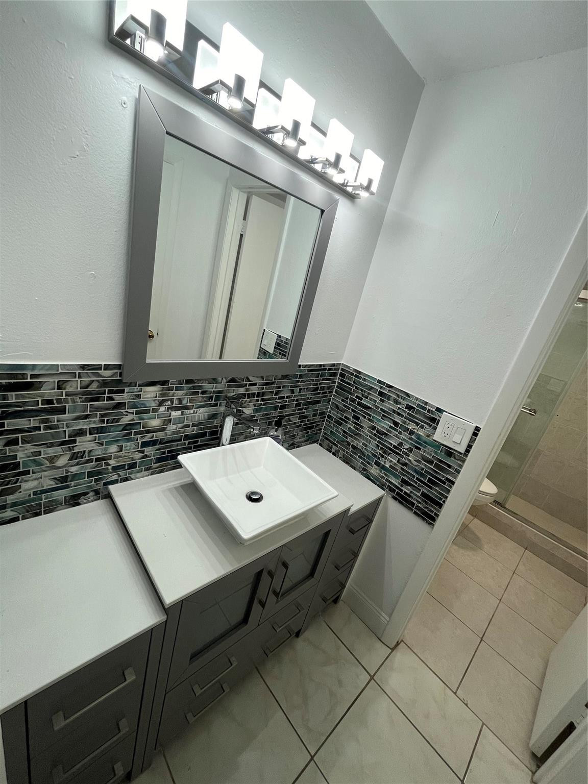 Bathroom vanity with a white vessel sink, dark mosaic backsplash, and large mirror.