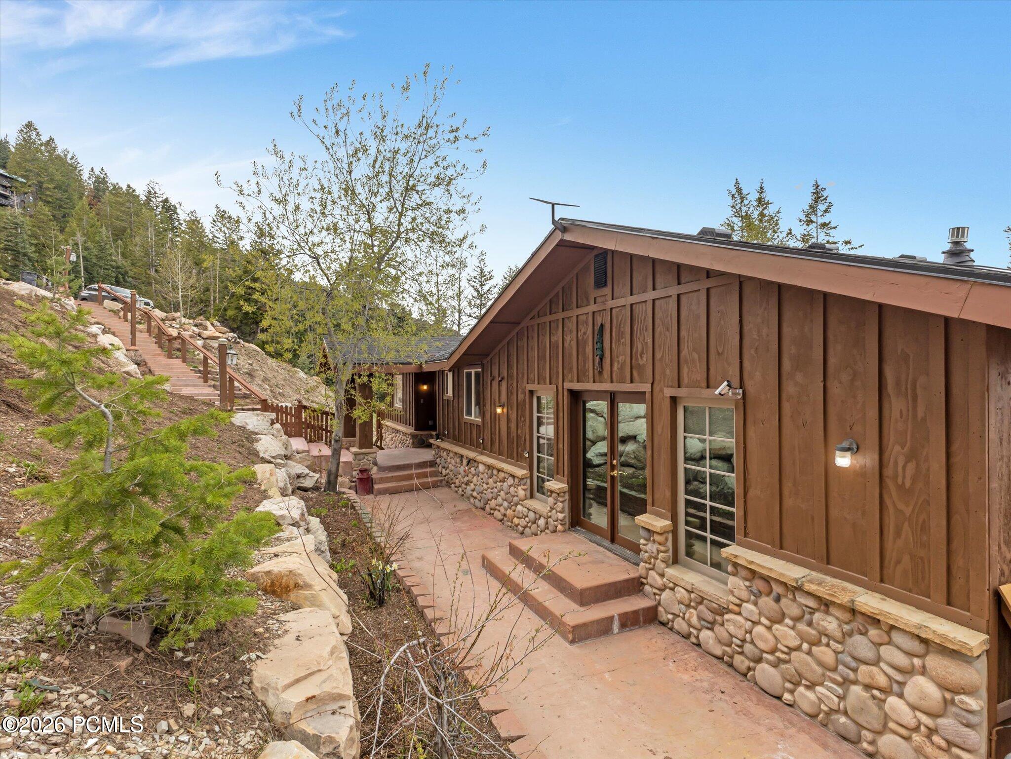 Brown wood-sided house with a river-rock foundation, beside a paved walkway and hillside steps.