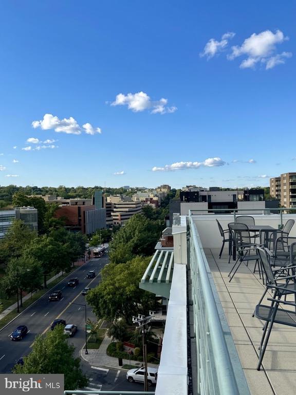 Rooftop terrace with glass railing and outdoor dining set overlooking a tree-lined city street.