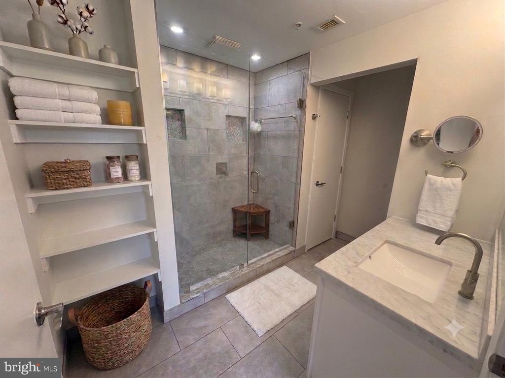 Bathroom with a glass-enclosed tiled shower, marble vanity, and open shelving with folded towels.