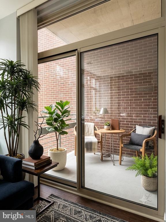 Condo interior with sliding glass doors opening onto a brick-walled balcony with seating and plants.