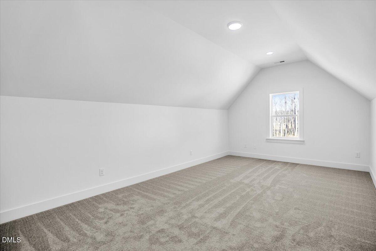 Empty carpeted upper-level room with a vaulted ceiling, recessed lights, and a window overlooking trees.