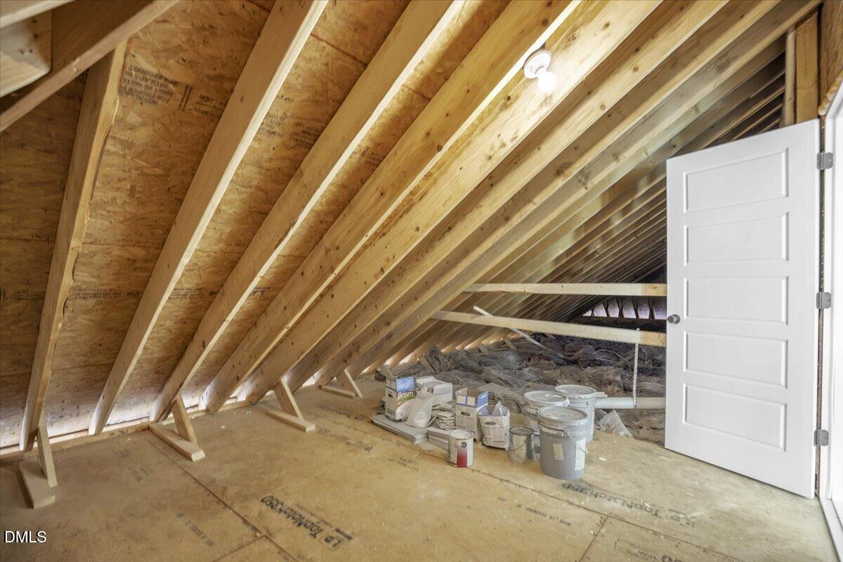 Unfinished attic with exposed rafters, plywood flooring, and paint supplies beside an open door.