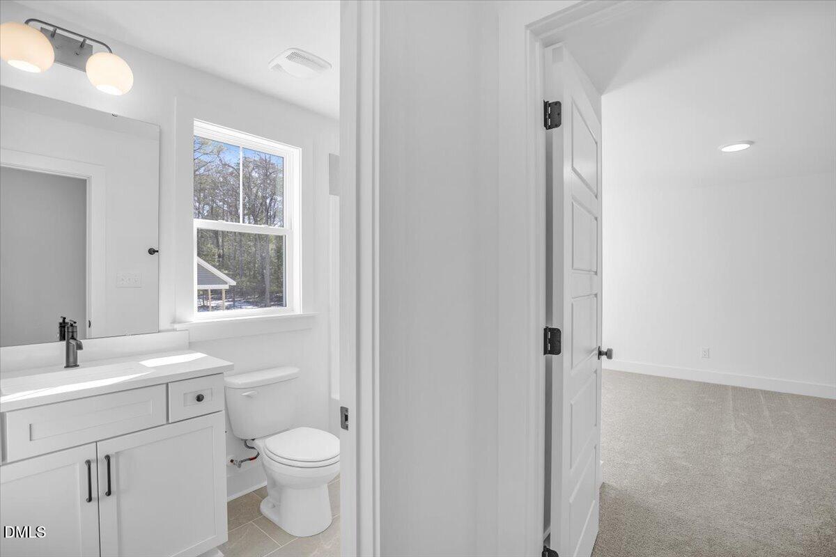 Bathroom with white vanity and toilet beside an open doorway to an empty carpeted room.