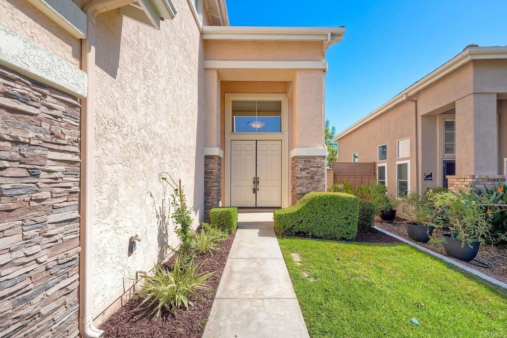 Stucco home entrance with stone accents, double doors, and a walkway bordered by lawn and shrubs.