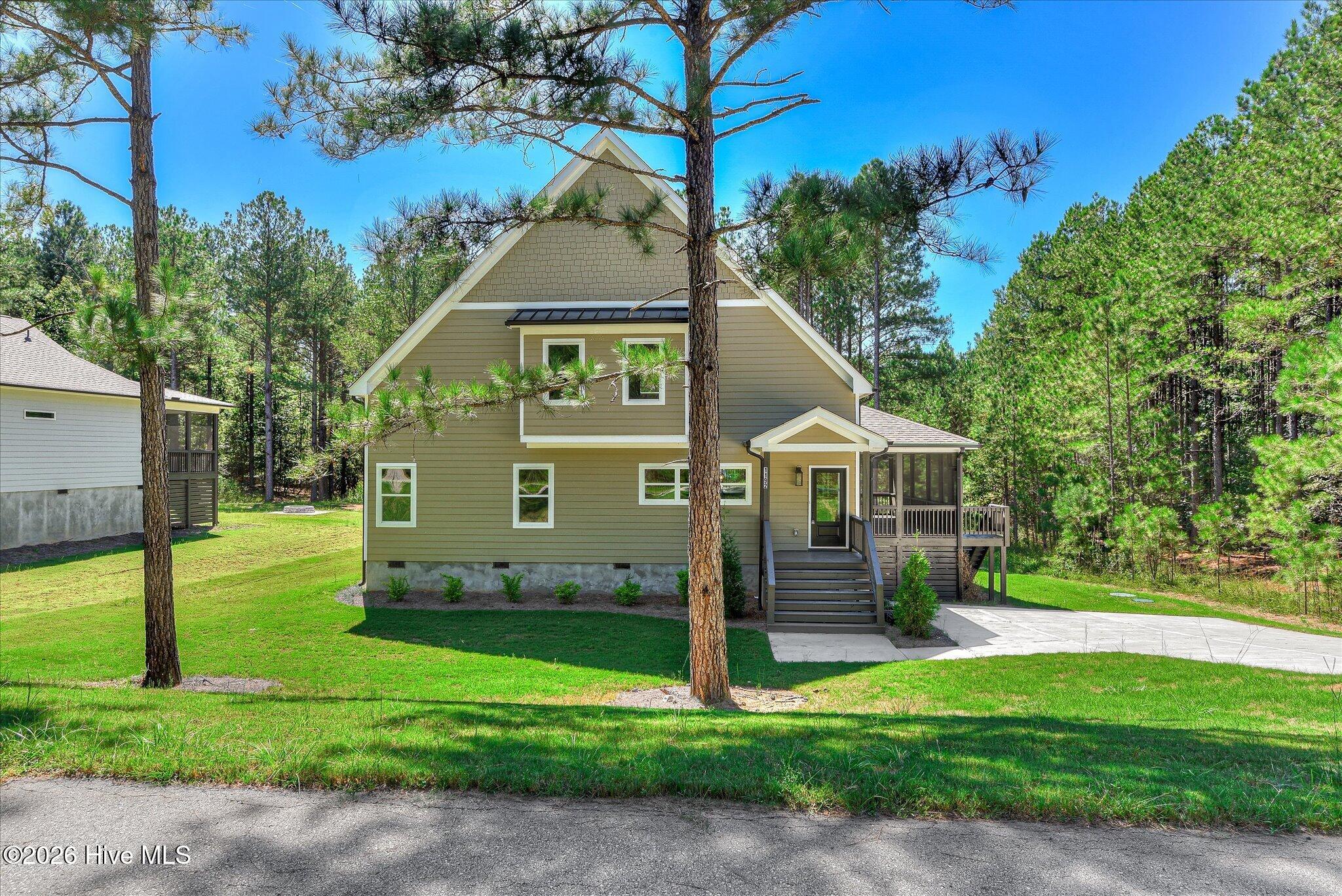 Two-story tan home with a steep gable and screened porch among pine trees.