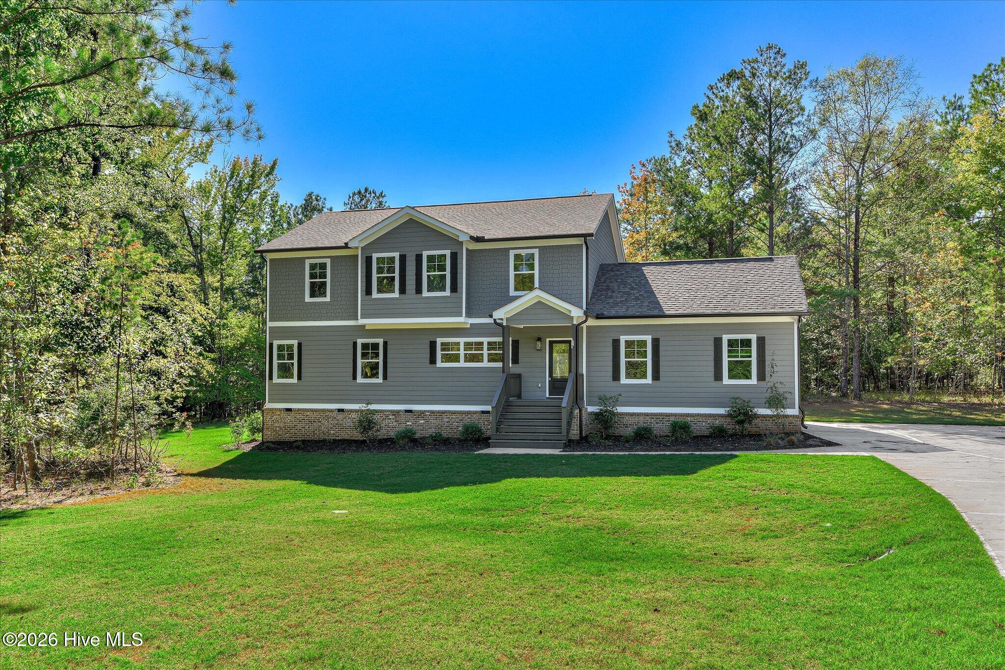 Two-story gray house with a covered entry, broad lawn, driveway, and mature trees.