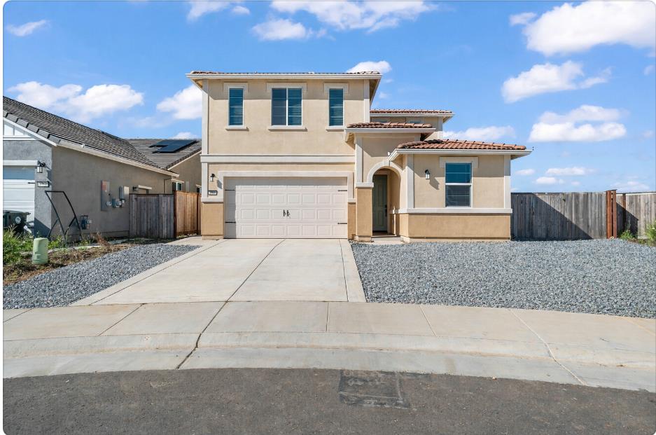 Two-story beige stucco house with an attached garage, wide driveway, and gravel landscaping.