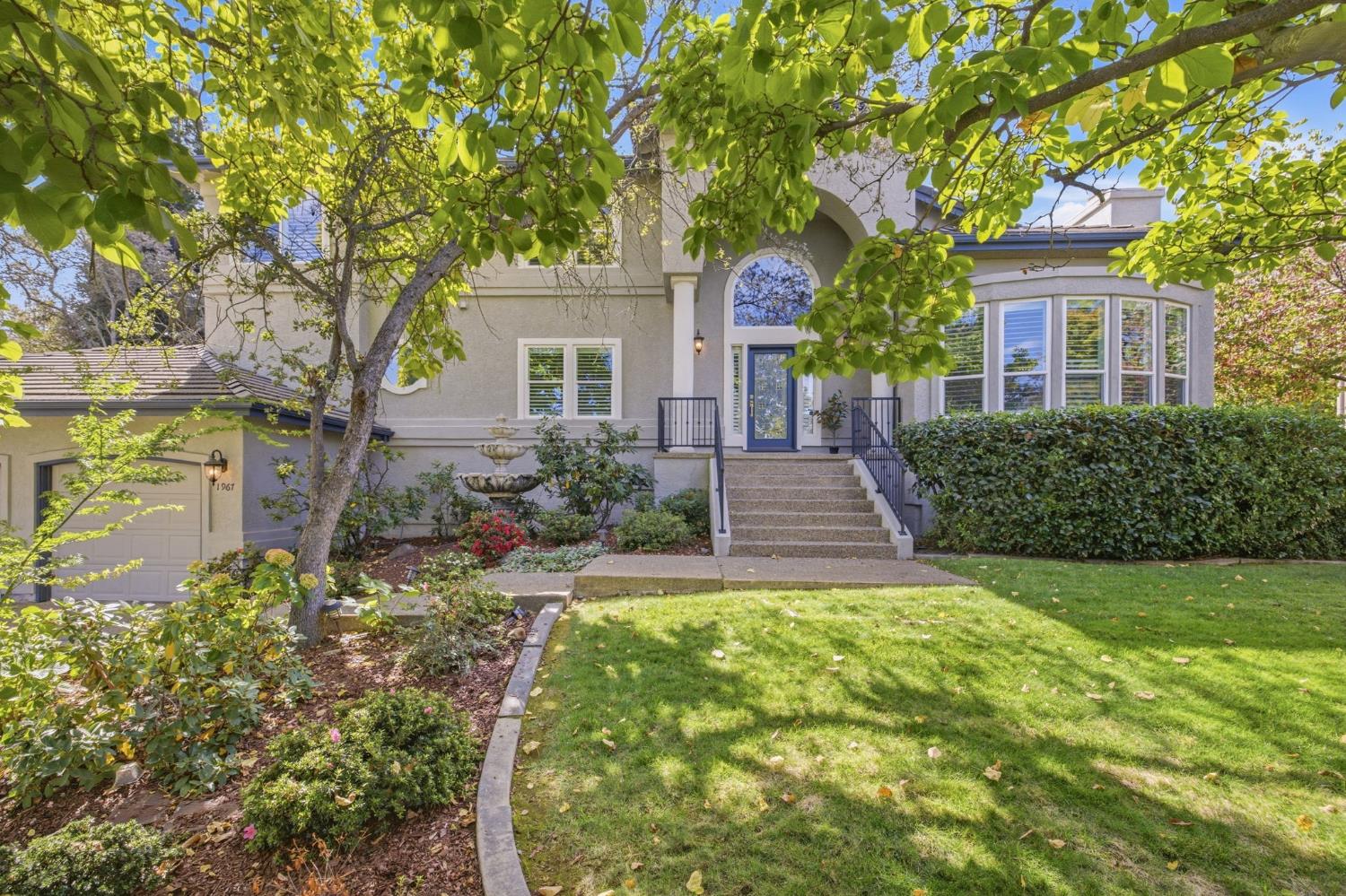 Gray two-story home with an arched entry, broad steps, bay windows, and a leafy front garden.