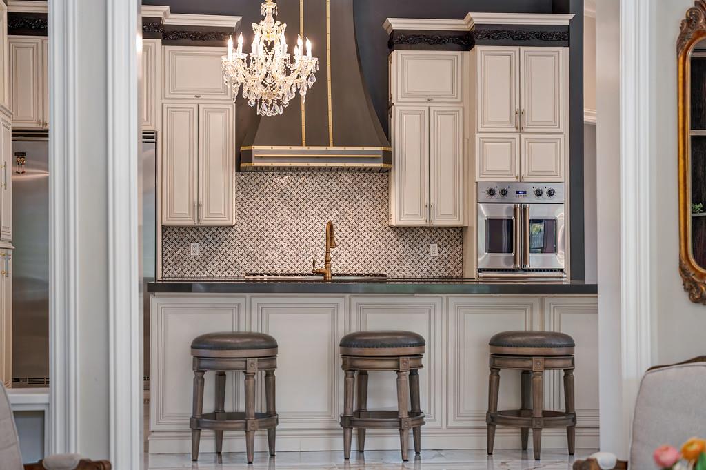 Kitchen with white cabinetry, patterned tile backsplash, three island stools, and a crystal chandelier.
