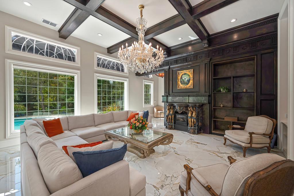 Living room with a crystal chandelier, dark wood fireplace, marble floors, and windows overlooking a pool.