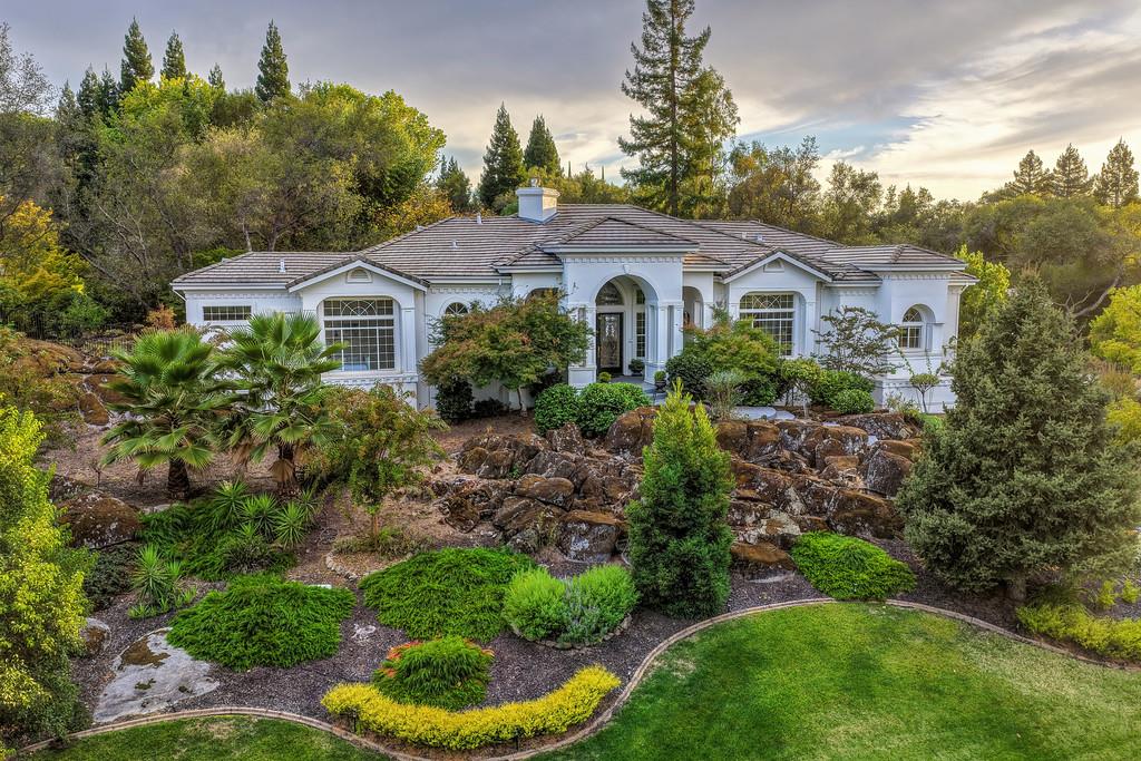 White single-story home with an arched entry, rock garden, and mature trees.