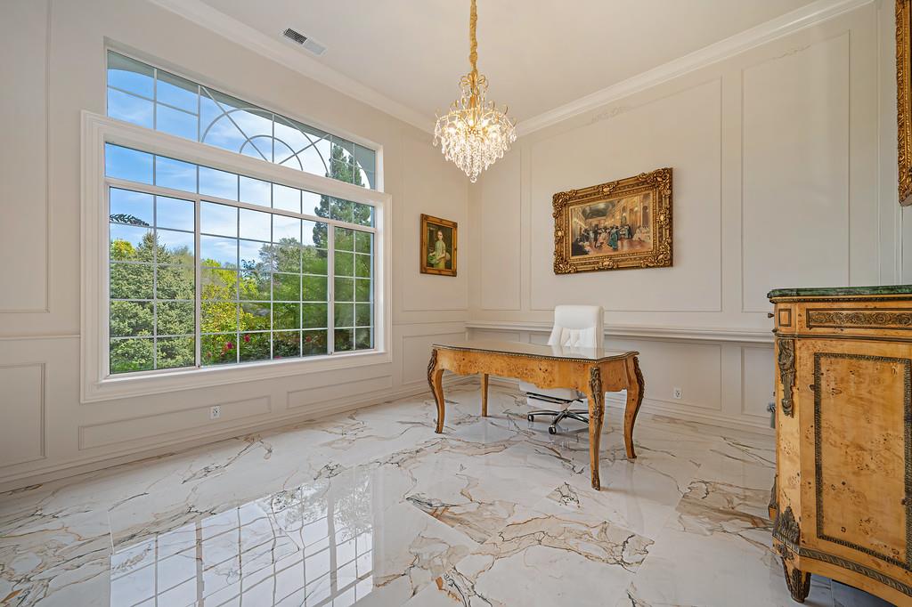 Bright office with a crystal chandelier, ornate desk, marble floors, and large window overlooking trees.