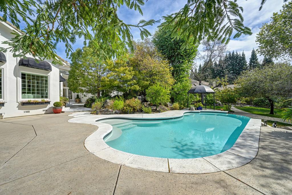 Curved swimming pool and stone patio beside a white stucco home, surrounded by mature trees and landscaped gardens.