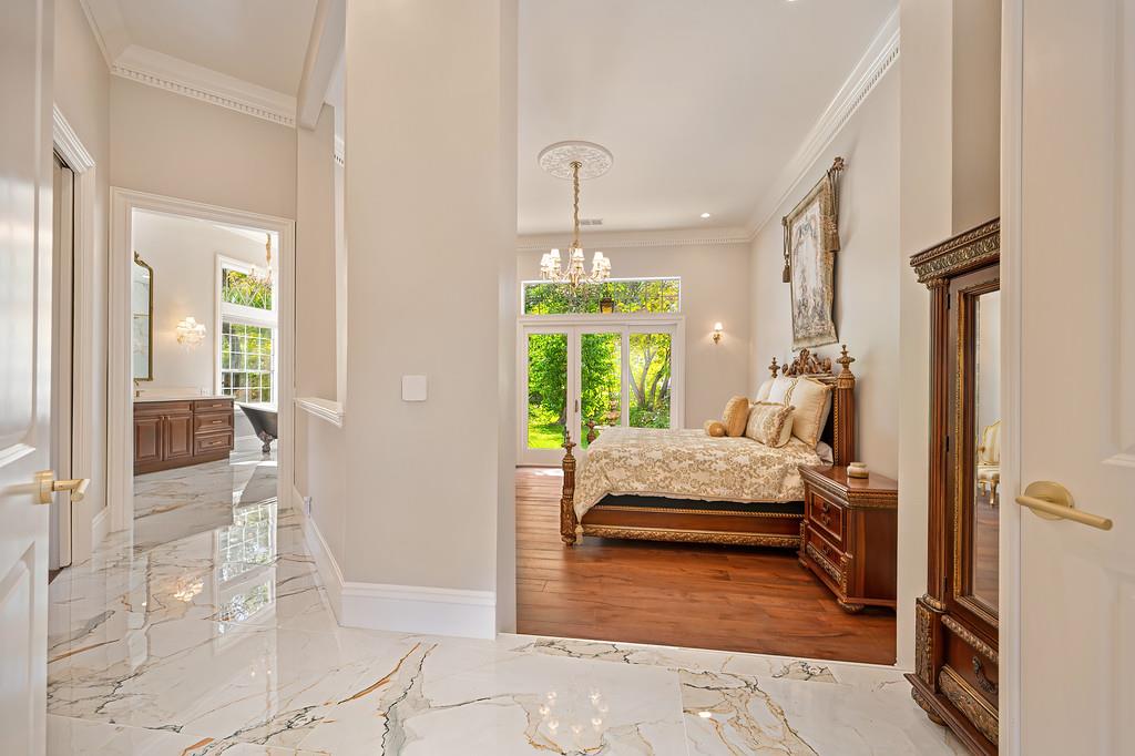 Furnished bedroom with a carved wooden bed and glass doors opening onto greenery, seen from a marble hallway.