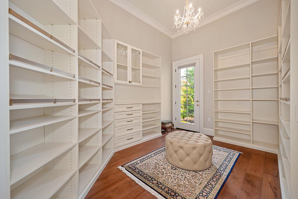Walk-in closet with white built-in shelving, a chandelier, and a tufted ottoman on a patterned rug.
