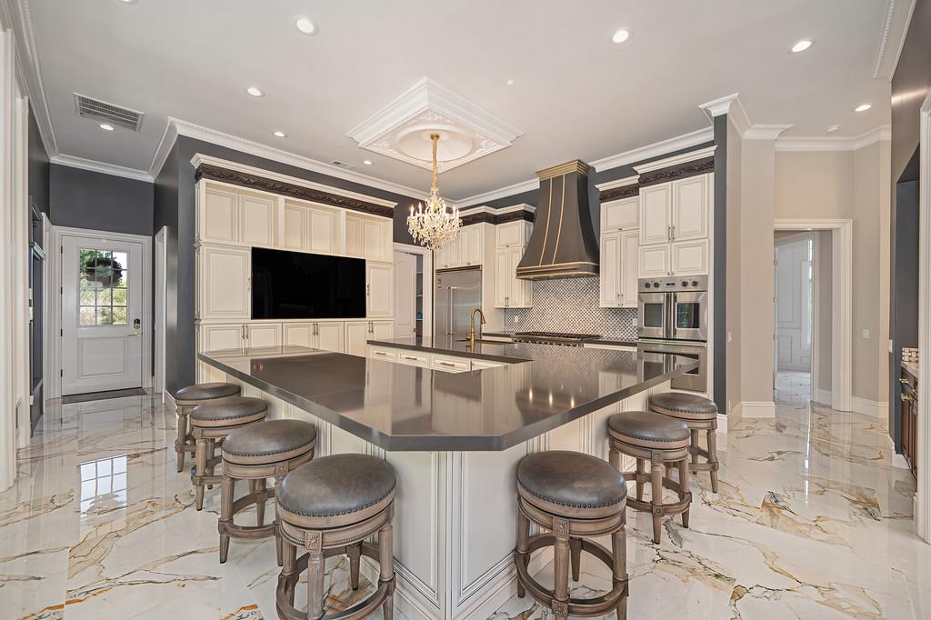 Ornate kitchen with a large island, upholstered stools, decorative range hood, and marble floors.