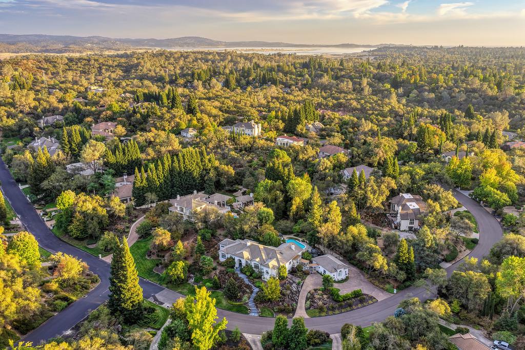Aerial view of a landscaped home with a pool, wooded neighborhood, and distant water.